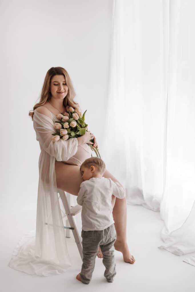 Schwangere Frau sitzt auf einer Leiter, mit Blumen in der Hand. Ihr Sohn umarmt ihre Beine