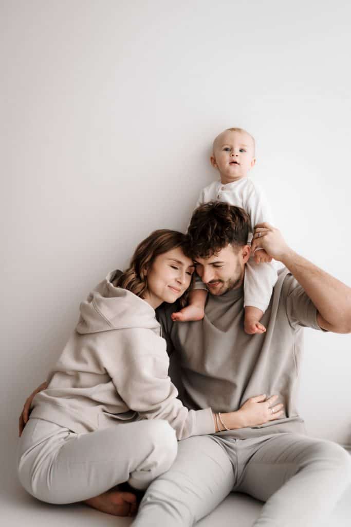 Eltern sitzen kuschelnd auf dem Boden, ein Kleinkind sitzt bei dem Vater auf der Schulter und schaut neugierig in die Kamera