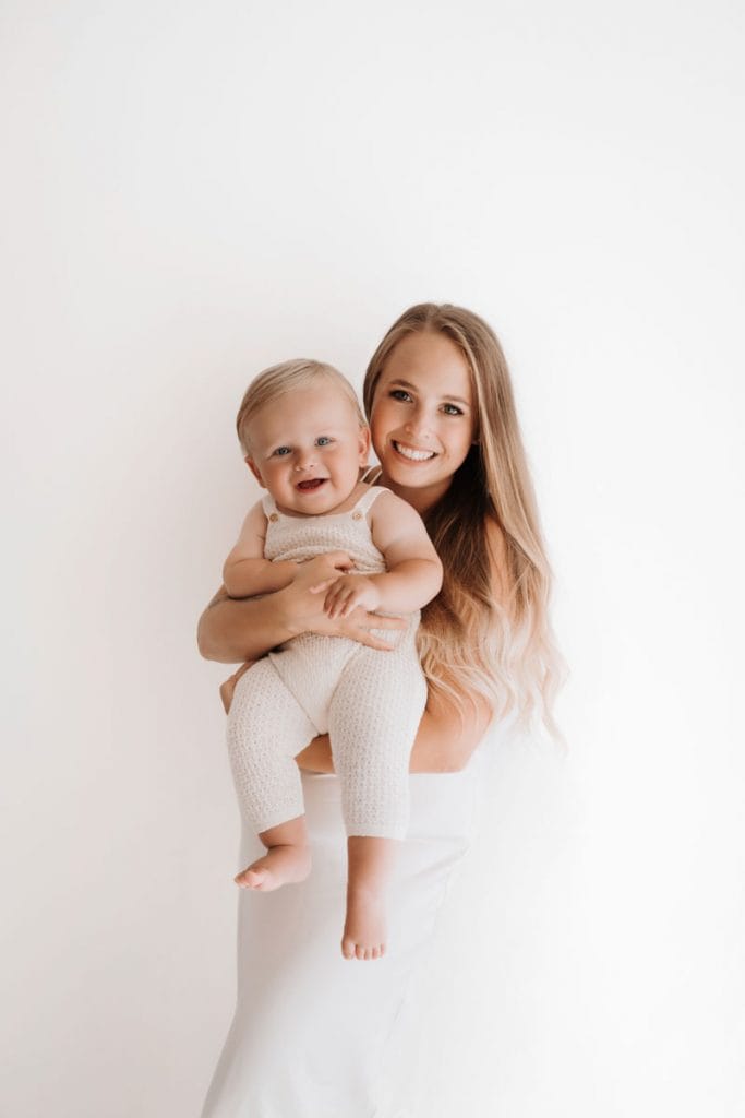 Mutter und Kind sind in hell gekleidet und stehen vor einer weißen Wand. Die Mama hält das Kleinkind auf dem Arm vor dem Bauch und beide lachen in die Kamera