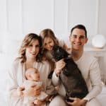 Junge Familie bestehend aus Mama, Papa, großer Tochter, Baby und Hund sitzen fröhlich lächelnd auf dem Bett.