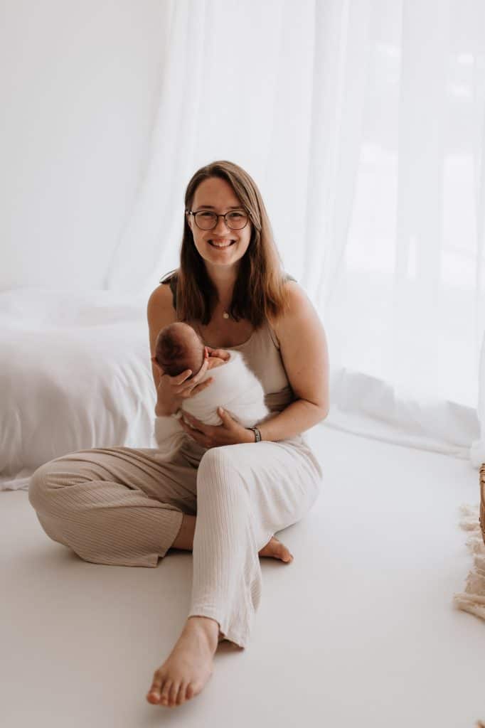 Farbige Fotografie einer Frau, die barfuß auf dem Boden sitzt und ein neugeborenes Baby in den Armen hält. Sie trägt ein bequemes, helles Outfit und lächelt herzlich in die Kamera. Der Raum ist hell und minimalistisch gestaltet, mit weißen Vorhängen und einem Bett im Hintergrund.