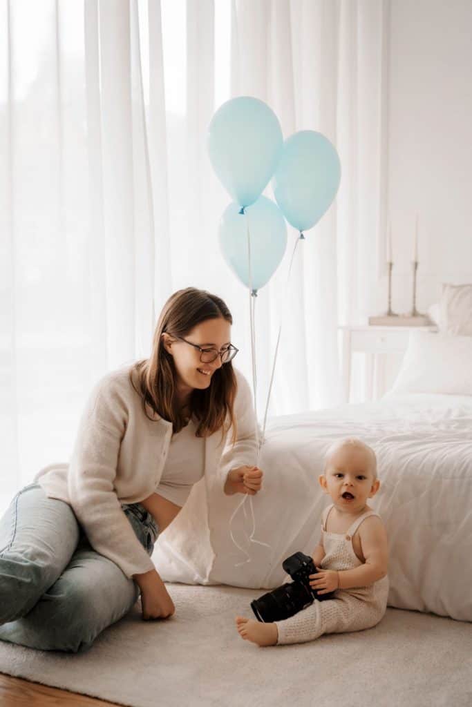 Ein Baby sitzt auf einem hellen Teppich in einem minimalistisch eingerichteten Raum und hält eine Kamera in den Händen. Neben ihm kniet eine lächelnde Frau mit einer Brille und hält hellblaue Luftballons. Im Hintergrund ist ein Bett mit weißer Bettwäsche zu sehen.