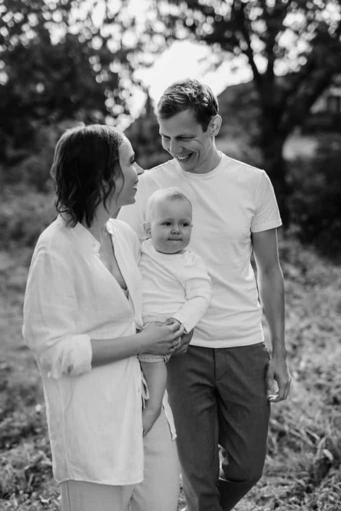 Ein Schwarz-Weiß-Familienporträt im Freien. Die Mutter hält das Baby im Arm, während der Vater sie liebevoll anschaut. Die Mutter und der Vater lächeln sich herzlich an, das Baby schaut ruhig in die Kamera. Im Hintergrund sind verschwommene Bäume und Lichtspiel zu erkennen, die eine warme Atmosphäre schaffen.