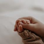Eine zarte Babyhand greift nach den Fingern einer erwachsenen Hand. Das Baby trägt einen gestrickten, braunen Ärmel, während die Haut der erwachsenen Hand sanft die kleine Hand umschließt.
