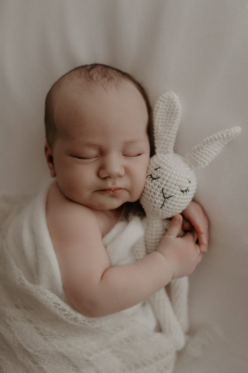Ein schlafendes Baby liegt eingehüllt in ein weiches Tuch und hält einen kleinen Stoffhasen im Arm. Die zarten Finger umschließen das Kuscheltier, während das Gesicht entspannt und friedlich wirkt.