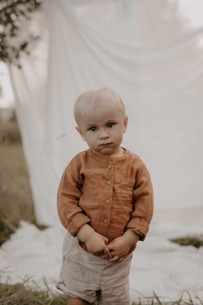 Ein kleiner Junge steht bei einem Outdoor-Shooting auf einer Wiese vor einem weißen Laken. Mit schüchternem Blick schaut er in die Kamera, während er seine Hände mit überkreuzten Fingern vor dem Bauch hält. Er trägt niedliche Leinenkleidung, die seinem natürlichen Look eine sanfte, zeitlose Note verleiht.