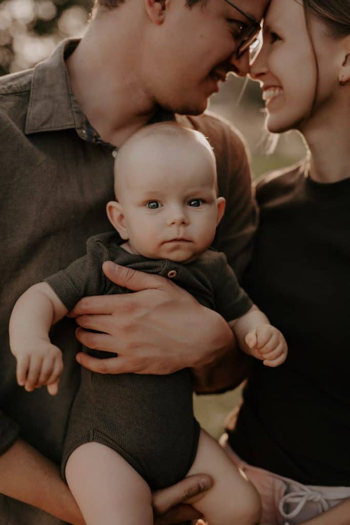 Im Hintergrund stehen die Eltern, Stirn an Stirn, und lächeln sich liebevoll an. Im Vordergrund hält der Vater das Baby auf dem Arm, das mit wachem Blick direkt in die Kamera schaut. Die warme Sonne scheint sanft durch die Bäume und verleiht dem Outdoor-Shooting eine harmonische, natürliche Atmosphäre.