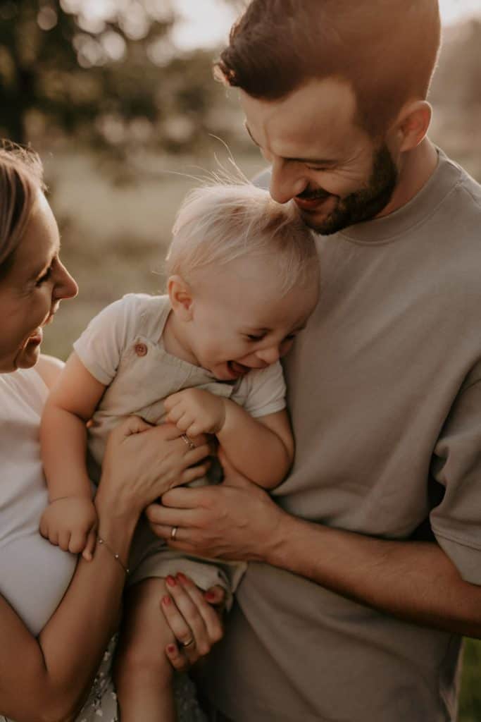 Mama und Papa halten ihr Kleinkind gemeinsam auf dem Arm. Die Mutter kitzelt es sanft, während es lachend an Papas Brust schmiegt. Beide Eltern schauen ihr Kind voller Liebe an. Im Hintergrund sind eine Wiese und Bäume verschwommen zu erkennen, was dem Outdoor-Shooting eine warme, natürliche Atmosphäre verleiht.