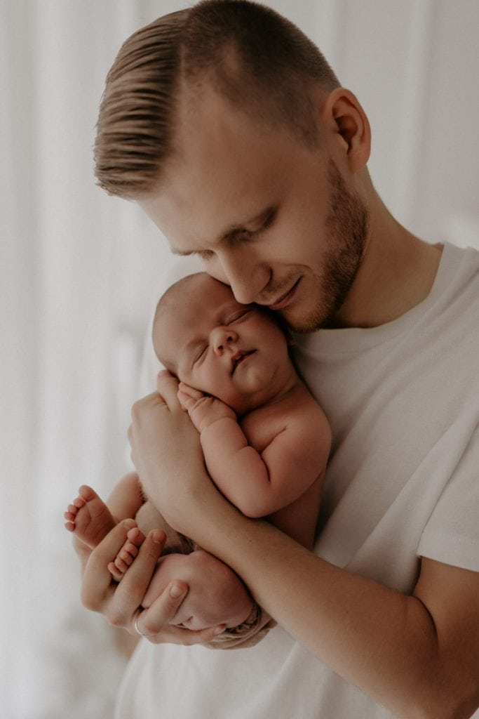 Ein Vater hält sein Neugeborenes mit beiden Händen liebevoll an sein Gesicht. Mit geschlossenen Augen schmiegt er sich sanft an das Köpfchen seines schlafenden Babys, während es friedlich in seinen Armen ruht.