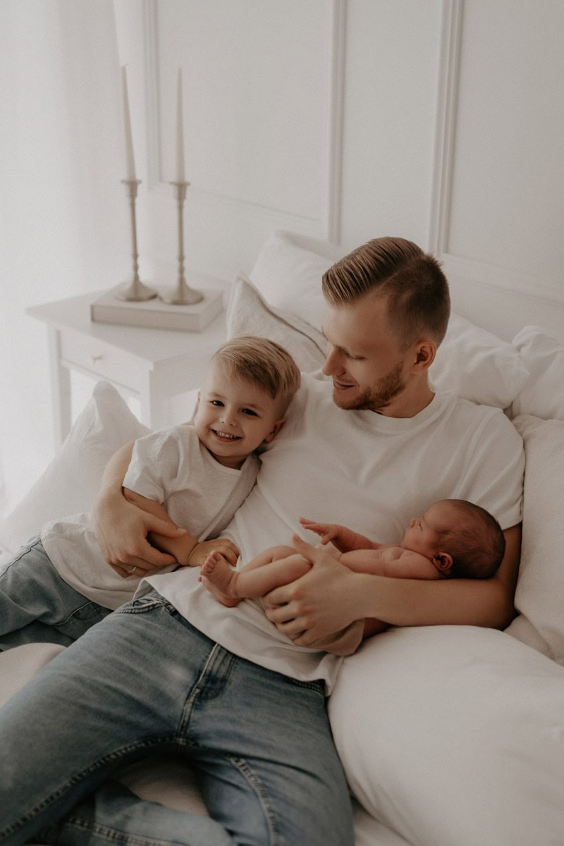 Ein Vater sitzt auf einem Bett und hält sein friedlich schlafendes Neugeborenes in einem Arm. Im anderen Arm sitzt sein größeres Kind, das fröhlich in die Kamera lächelt. Der Vater schaut es liebevoll an, während er beide Kinder behütet in seinen Armen hält.