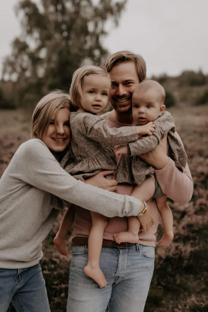 Eine glückliche Familie beim Outdoor-Shooting. Der Vater hält seine beiden kleinen Töchter liebevoll im Arm, während das älteste Kind sich lachend an ihn schmiegt. Alle tragen harmonisch aufeinander abgestimmte, natürliche Farben. Im Hintergrund sind eine Wiese und Bäume leicht verschwommen zu erkennen.