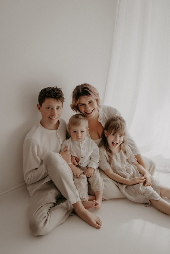 Eine glückliche Mama sitzt mit ihren drei Kindern in einem hellen, minimalistischen Raum vor einem weißen Vorhang. Sie trägt ein cremefarbenes Kleid und strahlt in die Kamera. Der ältere Sohn sitzt entspannt neben ihr, trägt ein beiges Shirt und eine helle Hose, und lächelt sanft. Das jüngste Kind sitzt auf ihrem Schoß, schaut ernst in die Kamera und trägt ein weißes Leinenoutfit. Die Tochter lehnt sich lachend an ihre Mama, trägt ein zartes Kleid und wirkt fröhlich und unbeschwert. Die warme, harmonische Farbgebung verleiht dem Bild eine weiche, liebevolle Atmosphäre.
