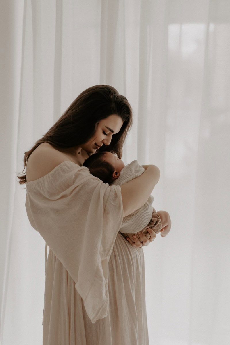 Eine Mama hält ihr Neugeborenes sanft in ihren Armen, während sie sich liebevoll zu ihm hinunterbeugt. Ihr Blick ist voller Wärme und Geborgenheit, das Baby kuschelt sich vertrauensvoll an ihre Brust. Sie trägt ein fließendes, beiges Kleid, das die zarte und innige Atmosphäre des Moments unterstreicht. Im Hintergrund weiche, helle Vorhänge, die das Bild in sanftes Licht tauchen.