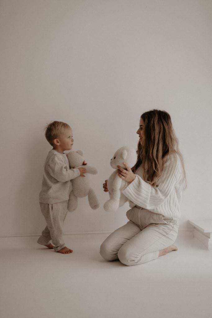 Ein kleiner Junge und seine Tante sitzen sich spielerisch gegenüber, beide halten einen weichen, cremefarbenen Teddybären in den Händen. Der Junge steht barfuß auf dem Boden und schaut seine Tante voller Freude an, während sie auf den Knien sitzt und ihn mit einem warmen Lächeln betrachtet. Ihre helle, gemütliche Kleidung harmoniert mit dem schlichten, ruhigen Hintergrund und unterstreicht die liebevolle Verbindung zwischen den beiden.