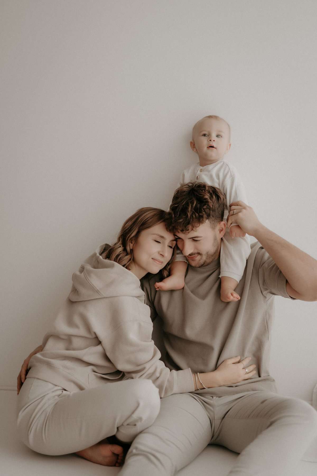 Ein junges Elternpaar sitzt eng aneinandergelehnt auf dem Boden, während der Vater ihr Baby auf seinen Schultern trägt. Die Mutter schmiegt sich mit geschlossenen Augen an ihn, während das Baby neugierig in die Kamera blickt.