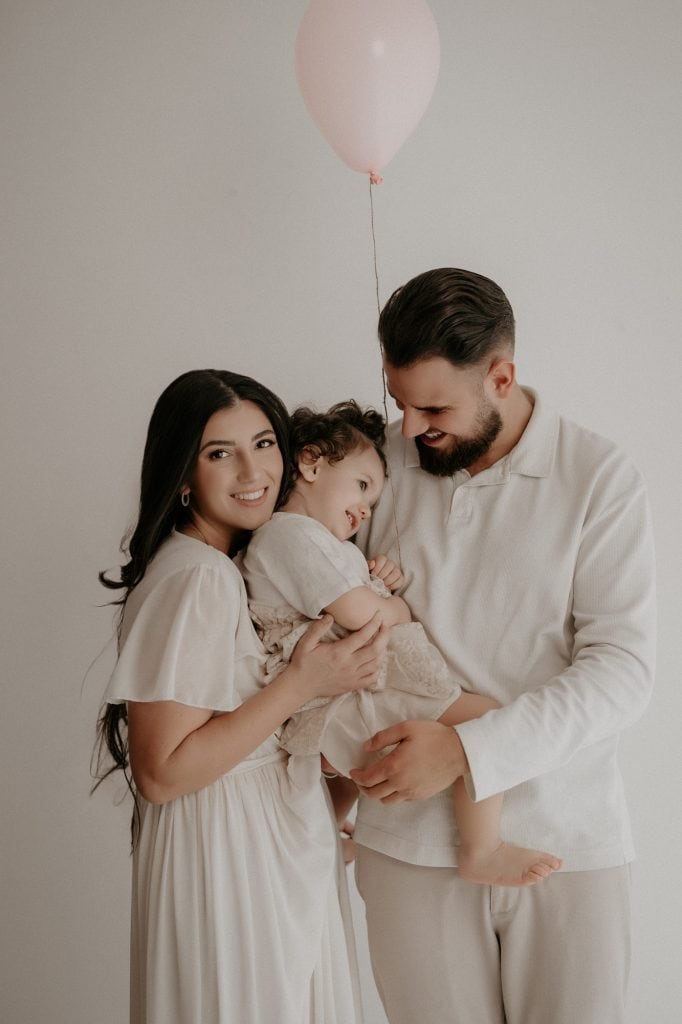 Ein Papa hält seine Tochter liebevoll auf dem Arm, während die Mama sich sanft von hinten an sie schmiegt. Die kleine Tochter lehnt sich entspannt an ihre Mama zurück und hält einen rosafarbenen Luftballon in der Hand. Der Papa blickt lächelnd seine Tochter an, während die Mama mit einem warmen Lächeln in die Kamera schaut