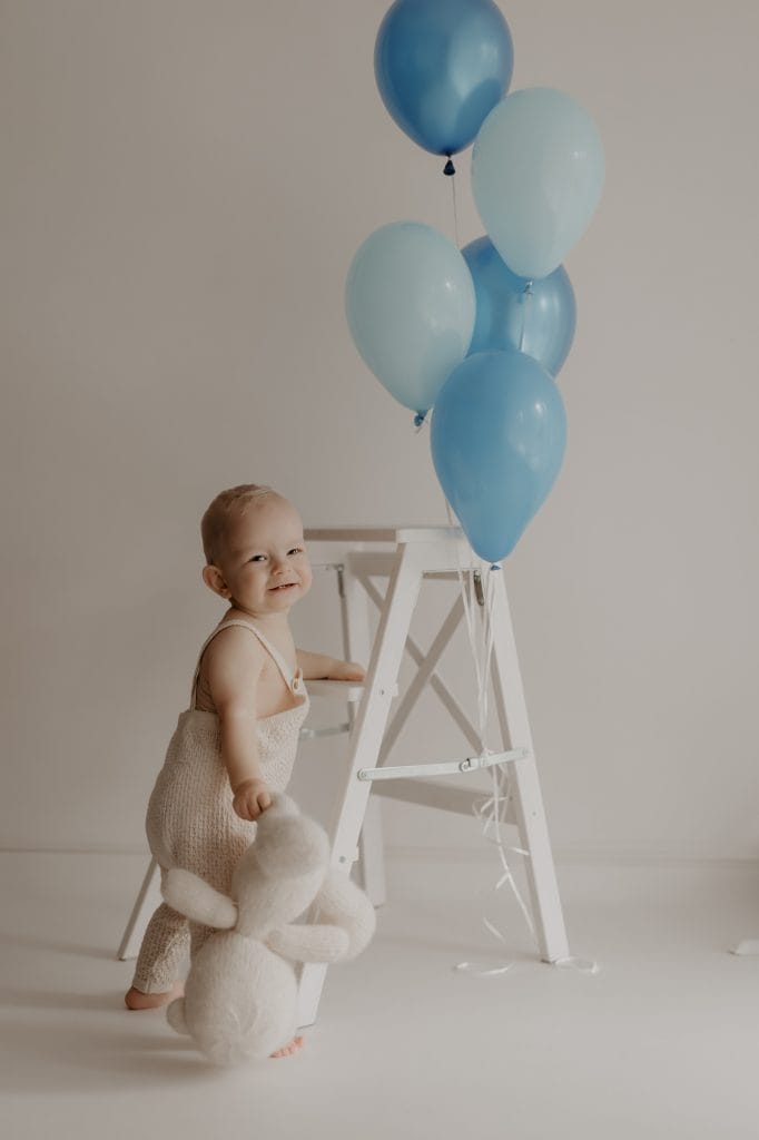 Ein kleiner Junge hält einen kuscheligen Teddy in der Hand und greift mit der anderen Hand nach einer weißen Leiter. An der Leiter schweben mehrere blaue Heliumluftballons. Der Junge dreht sich fröhlich zur Seite und blickt mit einem strahlenden Lächeln in die Kamera. Die Szene wirkt leicht und verspielt, mit sanften, neutralen Farbtönen im Hintergrund.