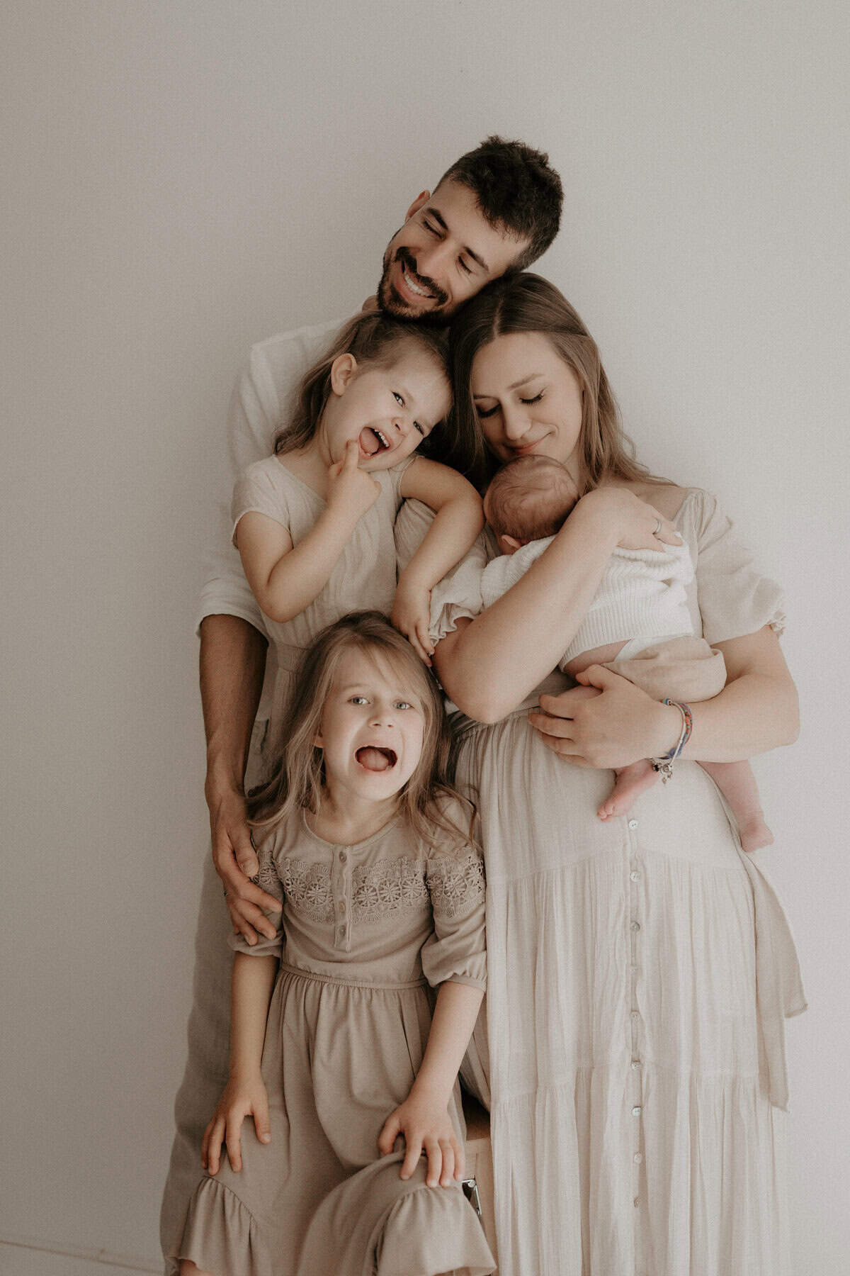 Eine junge Familie steht eng beisammen – Mama und Papa in der Mitte, während die beiden größeren Töchter lachend witzige Gesichter machen. Die Mutter hält das neugeborene Geschwisterchen liebevoll im Arm. Alle tragen beige Kleidung, was ein harmonisches und warmes Gesamtbild erzeugt.