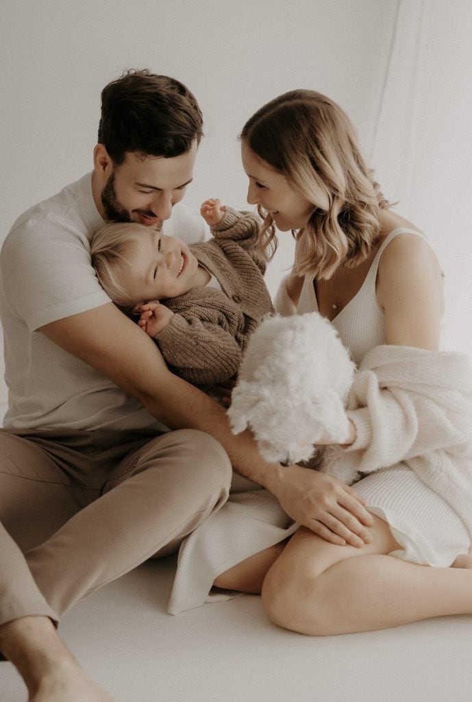 Junge Familie sitzt eng beisammen auf dem Boden. Der Papa auf der linken Seite, die Mama auf der rechten, in der Mitte ihr kleiner Sohn, der sich lachend an die Brust seines Papas kuschelt. Die Mama hält einen kleinen Hund auf dem Schoß und blickt ihren Sohn liebevoll an. Beide Eltern strahlen Wärme und Geborgenheit aus, während sie ihren fröhlichen Sohn ansehen.